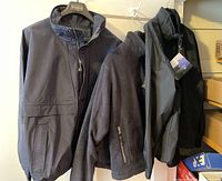 Three men's jackets hung together; black windbreaker, dark gray fleece, and black shell with visible logos and tags.
