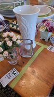 Photo of three vases on wooden table: white porcelain Kaiser vase, small signed crystal vase, and a small glass vase with artificial flowers
