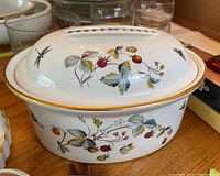 Porcelain roasting pan with matching lid showing Strawberry Fair design with strawberries, leaves, small flowers and insects like dragonflies and butterflies. Gold trim along the edges. Oval shaped with integrated lid handle.