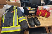 Overall lot on table showing Pioneer safety jacket with reflective stripes, black Dakota safety shoes, two bags with survival and emergency road kit items, and white gloves.