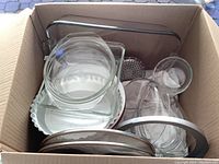 Box containing various clear glass kitchenware such as bowls, baking dishes, glass pie dish, and metal trays with a mesh strainer.