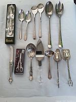 Overview of all assorted vintage silverplated serving cutlery items arranged on white cloth including spoons, forks, tongs, a ladle, cake server, scissors, and serving knife, some boxed.