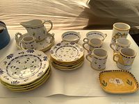 Wide shot showing the entire set of hand painted Italian ceramic dishware arranged on a table, including plates, mugs, cups, creamer, sugar bowl, and a serving dish.