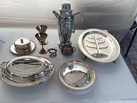 Full view of entire silverplated servingware lot arranged on white foldable table, showing all pieces and overall condition with some tarnishing.