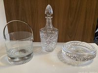 Photo showing Waterford crystal ice bucket with metal handle, crystal decanter with stopper, and crystal ashtray on white surface against wooden backdrop.