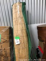 Rolled bamboo blind with green protective cover, standing vertically next to cardboard boxes in storage.