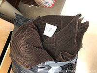 Brown quilted fabric bedspread folded and placed on a black surface, with a white lot number tag visible.