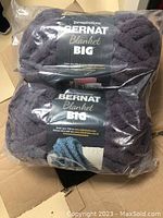 Eight balls of Bernat Blanket Big purple yarn packaged in plastic, placed in a cardboard box.