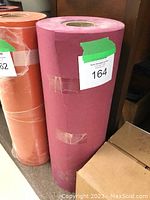 Large roll of rose-colored butcher paper standing vertically next to other rolls and a cardboard box, with tape marks and a label attached