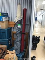 Two fabric rolls, one red upholstery fabric and one cream cheesecloth fabric, standing upright in storage.