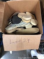 Photo of an open cardboard box filled with assorted large rubber shoe soles in black, beige, and brown colors showing different tread patterns and styles.