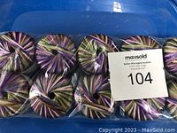 10 balls of Needful Yarns Elba yarn packed in a clear plastic bag, showing variegated colors in purple, green, yellow and pink.