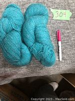 Two skeins of blue wool yarn placed on a grey fabric surface with a marker beside for size reference and a note marked 304 nearby.