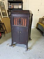 Full view of the closed antique gramophone console showing dark wood cabinet with carved Gothic style front panel, brass knobs, and decorative feet on casters.
