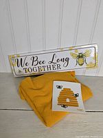 Photo of all three bee themed items: metal sign, yellow throw, and Swedish dishcloth arranged on a wooden surface with white wall background
