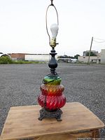 Photo of a multicolored swirled glass base lamp with a black metal neck, ornate metal base, and visible light bulb socket with harp.
