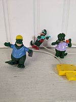 Three vintage Dinosaurs TV show Happy Meal toy figures with varying costumes and accessories, displayed upright on white background. Yellow controller with cord visible.