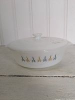 Side view of Fireking white milk glass baking dish with black and gold starburst pattern, showing handle and lid knob