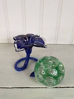 Blue glass flower-shaped vase with clear edges and green glass paperweight positioned on a white surface.