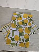 Two folded dish towels; one lemon print, one yellow-green-white plaid, displayed on a gray wooden surface under white paneled background