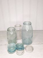 Three vintage mason jars of varying sizes with embossed text on glass, along with five additional glass jar lids mostly blue, one clear.