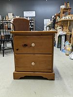 Front view of the pine wood bedside table showing two drawers with white knobs and carved base.