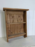 Front view of unfinished pine wall shelf with two shelves, showing vertical plank back panel and a slightly decorative bottom edge on sides.