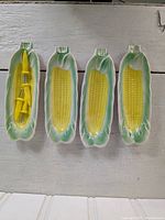 Four ceramic corn holders shaped and painted to resemble ears of corn with yellow kernels and green husks, two with yellow plastic corn holders inside.