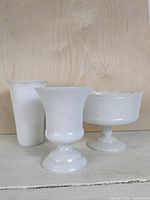 Photo of three white opaque milk glass items set against a wood background. Items include a tall cylindrical vase, a pedestal flared rim vase, and a pedestal bowl with scalloped edge.