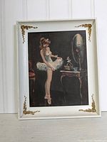 Photo of framed ballerina art print showing the ballerina looking in a mirror, with white plastic frame and gold decorative corners.