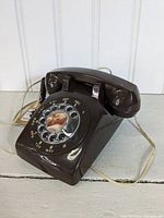Front angled view of vintage brown rotary dial phone showing rotary dial, handset, coiled cord, and beige wiring cable.