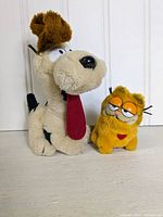 Full view of both vintage plush toys showing Odie on the left and Garfield on the right, displayed standing upright on a wooden surface against a white paneled wall.