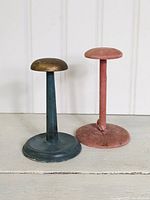 Two vintage hat stands on a white surface against a white paneled background, one wooden and blue, one pink velvet.
