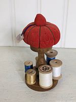 Front view of vintage red tomato-shaped pin cushion on wooden holder, surrounded by various thread spools in colors such as white, gold, and blue.