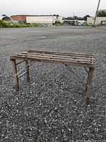 Photo of antique wooden wash stand outdoors on gravel, showing front and side view with slatted top and foldable legs.