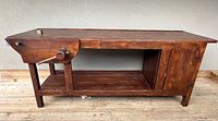 Full side view of the antique carpenters bench showing the solid wood construction, wooden vise with handle, open storage shelf, and cabinet with hinged doors.