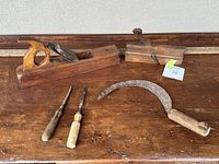 Full lot overview showing 2 wooden hand planes, 2 chisels, and a curved sickle-shaped tool, laid out on a wooden surface.