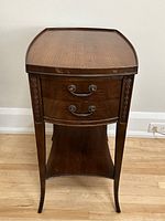 Front angle view of the mahogany antique end table showing curved drawers, metal handles, carved floral trim, and lower shelf.