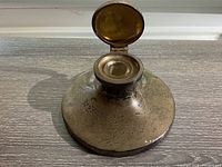 Top and side views of the antique sterling silver inkwell showing the hinged lid open revealing glass ink pot, the patina on the silver surface, and overall circular shape.