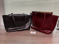 Two large handbags side by side, a red faux alligator and a black leather bag on table in front of window.