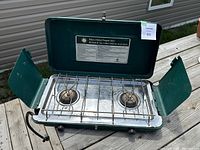 Open view of the green metal stove showing two burners, metal grill grates, control knobs, and fold-out wind guards.