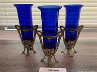 Front view of three 7-inch blue glass vases set in a decorative metal Art Nouveau style stand with detailed scroll and floral designs, showing the vases upright with flared lips.