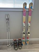 Full view of Roxy skis, Rossignol women's ski boots, and Kerma ski poles against a wall and window.