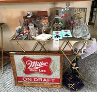 Overall view of barware items displayed on two glass-top folding tables with gold-colored legs. Items include various glasses, decanter, bottle openers, coasters, chip and dip bowl, a lamp on the floor, and the Miller High Life On Draft sign leaning against the tables.