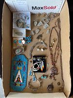 Top-down photo showing an assortment of costume jewellery including earrings, bracelets, necklaces, pendants, and an indigenous beaded art pouch inside a cardboard box.