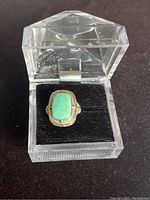 Gold ring with rectangular green stone in acrylic display box on black fabric background.
