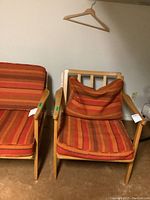 Front view of mid century modern wooden armchair next to matching chair, showing orange and red striped cushion with loose back cushion.