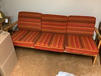 Full view of mid century modern sofa with three striped orange/red/brown cushions and wooden armrests, placed in corner of room with boxes nearby.