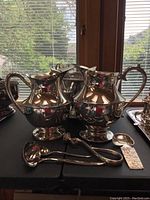 Photo showing two silver plated water jugs with curved handles and spouts placed on black surface.