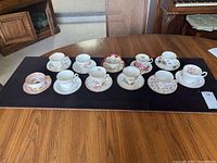 Top view of eleven teacups with matching saucers arranged on a black mat on a wood table, showing various floral and gold designs.